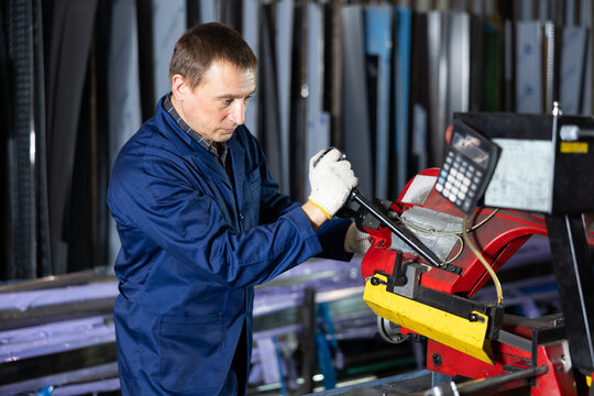 Serious experienced middle-aged metal worker in blue overalls and gloves operating pipe cutting machine in industrial workshop