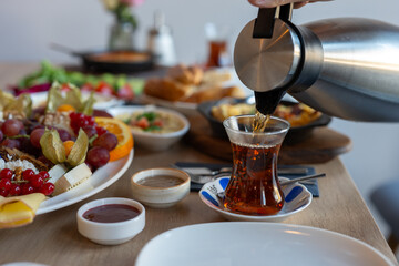 Fototapeta premium Pouring Turkish Tea From Kettle Into Glass Cup With Fruit Platter And Breakfast Dishes On Wooden Table For Food And Drink Concepts