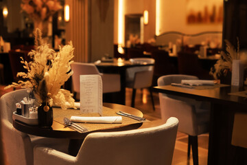 Elegant restaurant table setting with menu, cutlery and dried flowers in vase under warm ambient lighting.
