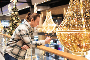 Young man leaning on railing using phone amidst festive Christmas lights and decorations in a shopping mall.