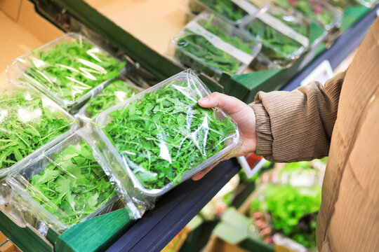 Customer holding a plastic container of fresh green arugula leaves while shopping in the supermarket produce section.