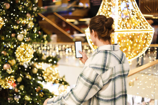 Man taking photo of Christmas decorations in a shopping mall using a smartphone with a blank white screen.