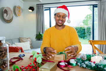 African American woman wearing Santa hat sitting at table tying green ribbon around brown gift box