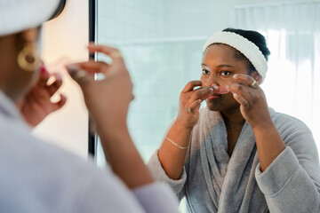African American woman applying translucent nose pore strip at bathroom mirror wearing bathrobe