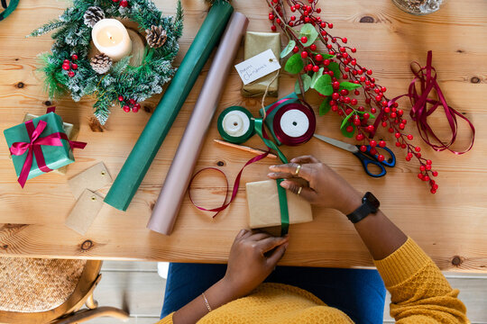 Small kraft package being tied with green ribbon on wooden tabletop, surrounded by festive wreath
