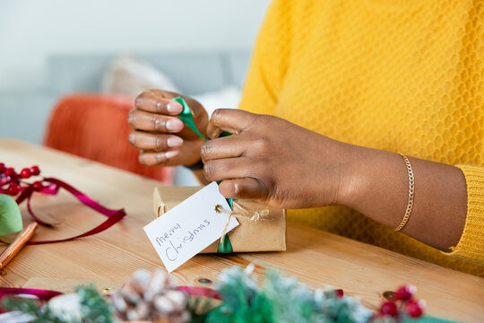 Hands tying green ribbon around brown craft paper gift on wooden table, with festive decorations