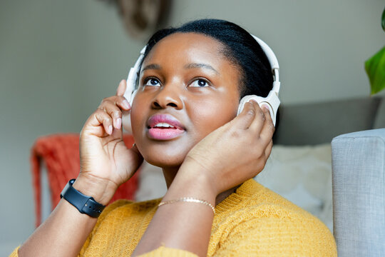 African American woman adjusting wireless headphones while wearing mustard sweater on sofa at home