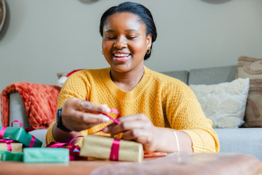 African American woman tying pink ribbon around gifts on wooden coffee table at home