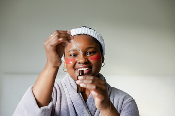 African American woman pipetting serum into bottle at home wearing headband red under eye patches