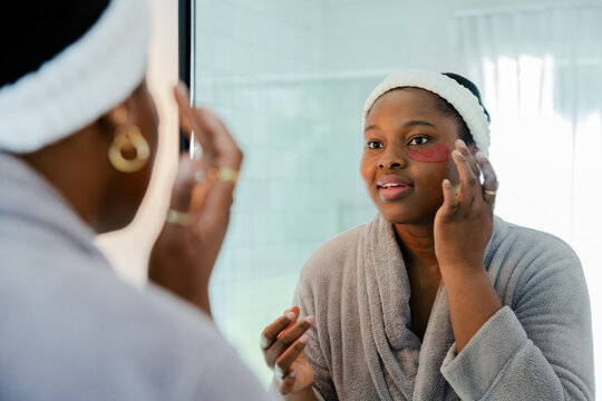 African American woman applying red under-eye gel patches at bathroom mirror wearing spa headband