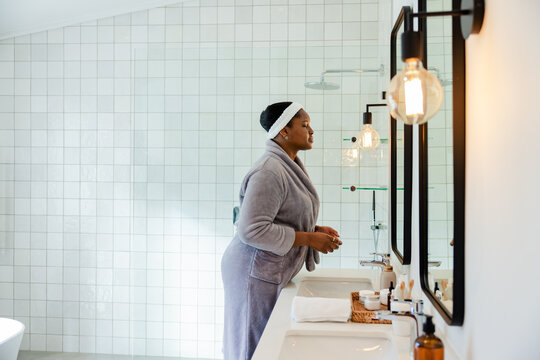 African American woman wearing robe, headband bending at vanity with skincare bottles, copy space