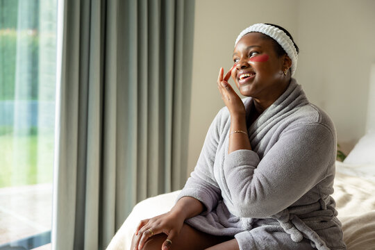 African American woman in grey bathrobe sitting on bed by window applying skincare cream to cheeks