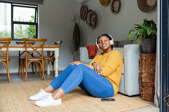 African American woman sitting on rug wearing sweater listening to music at home with smartphone