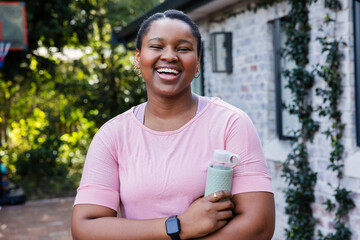 African American woman standing smiling on backyard patio holding water bottle, wearing smartwatch