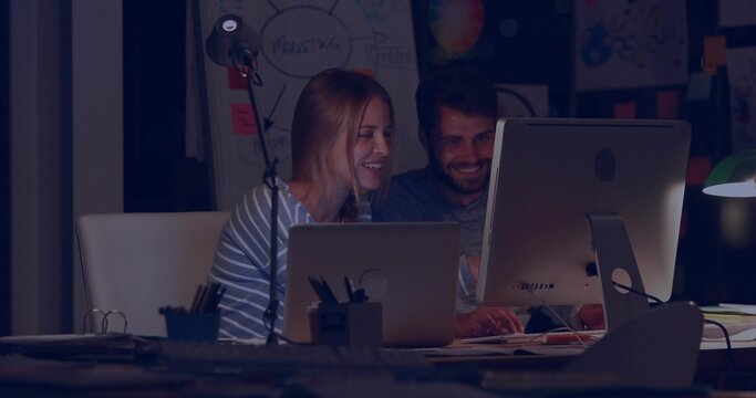 Leaning, pair wearing dark shirt, striped top reviewing code in dim workspace with monitor, laptop