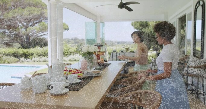 Arranging dishes, two women placing teacups on long counter on covered patio with teapot and fruit - Powered by Adobe
