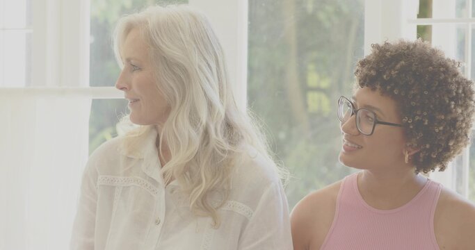 Talking senior and adult women sitting at home by window, wearing blouse and pink top, glasses
