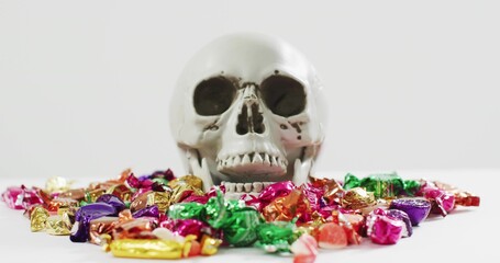 Fototapeta premium Resting skull replica sitting centered on white tabletop, surrounded by foil-wrapped candies