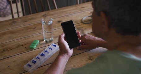 Holding smartphone, senior wearing teal shirt inspecting pill organizer at home table, copy space