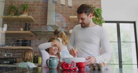 Stirring man and daughter mixing bowl on kitchen island, with red measuring cups and egg carton