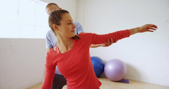 Stretching woman in red top doing side-arm stretch in rehab studio, trainer guiding with balls