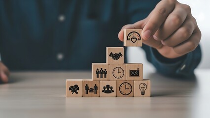 Man stacking wooden blocks with business symbols on a table, representing growth, success, and achievement in a focused and professional setting.