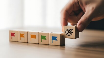Hand places wooden block with target symbol onto a row of blocks with flag symbols on a wooden table, representing goal achievement and strategic planning.