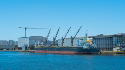 Large general cargo vessel moored at a busy commercial port terminal with dockside warehouses, a large crane, and an urban skyline