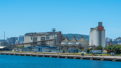 Obraz premium Large port facility featuring industrial grain silos, a tall loading tower, and massive storage buildings against a cityscape backdrop