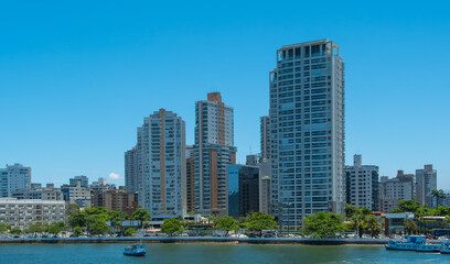 Skyscrapers and modern residential buildings line a sunny waterfront cityscape with clear blue skies and green trees, reflecting urban life