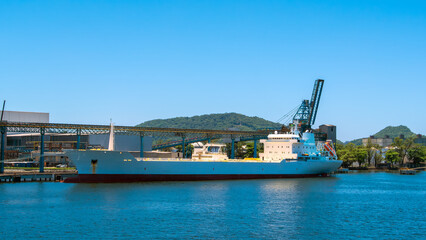 White cargo tanker ship docked in a busy port with industrial conveyor structures and a green, forested hill in the background