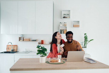 Diverse couple laughing, enjoying online shopping with credit card