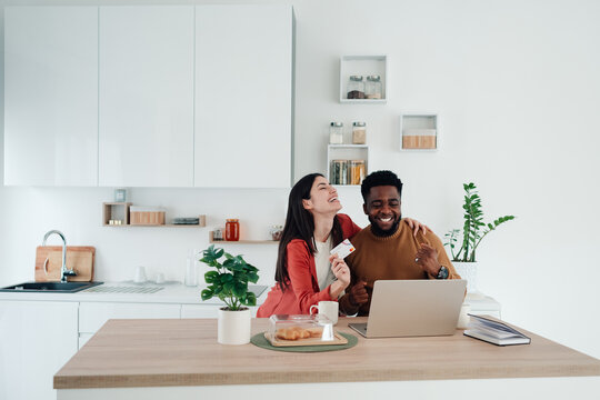 Diverse couple laughing, enjoying online shopping with credit card