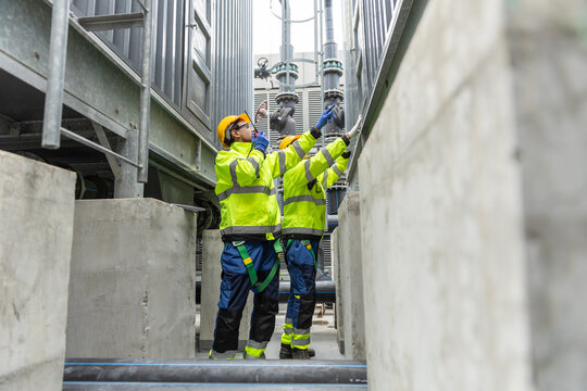 Two industrial engineers in safety uniform inspecting and discussing machinery at factory site. Concept of teamwork, maintenance, technical inspection, workplace safety in industrial environments.