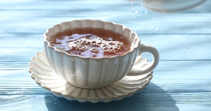 Pouring hot tea into cup from teapot at light blue wooden table, closeup