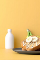 Close-up of toast with banana slices and white drizzle on a plate, with a bottle and bananas in the background, set against a yellow backdrop.