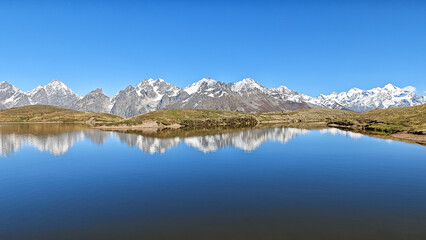 Crystal clear glacial lake reflecting the dramatic snow-capped peaks of the Caucasus Mountains and the background sky under a perfect blue sky near Mestia, Georgia