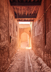 Narrow, dimly lit, dusty alleyway between tall, traditional mud-brick walls in a Moroccan village or old town, leading to a sunlit archway and open space