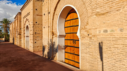 Long side view of the textured terracotta brick wall of a Marrakech mosque leading to a beautiful studded wooden door set in a classic Moorish arch 