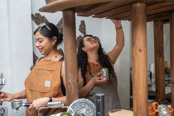 Two young women behind the counter preparing coffee together, focused and smiling while working in their café. Small business barista skills, coffee preparation, teamwork in a welcoming shop