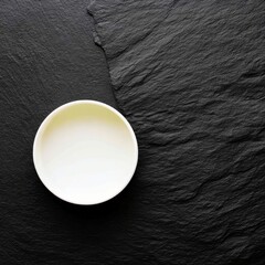 An overhead shot of an empty white bowl on a textured black slate stone surface. The bowl is centered, and the lighting is soft.