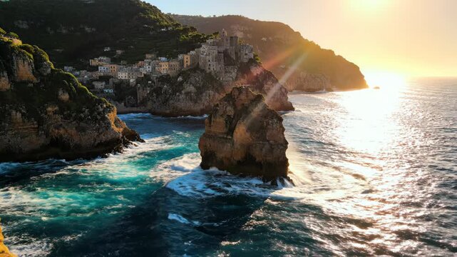 Coastal village perched on cliffside overlooking turquoise sea with dramatic wave crashing against sea stack at sunset 4k video