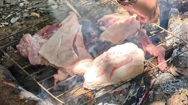 Footage of a man's arm placing raw meat on a simple outdoor grill over hot coals. Primitive open-fire cooking in nature with smoke and flames rising from the fire pit.