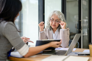 Old asian businesswoman holding glasses talking to employee office worker at table in company room.