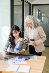 Old asian businesswoman holding coffee looking at folder helping a young coworker at office table