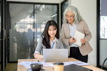 Old asian businesswoman standing holding pen and book looking at young coworker's laptop in office
