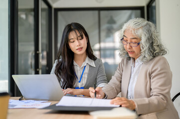 Obraz premium Old asian businesswoman holding pen explaining documents to young coworker while sitting in office.