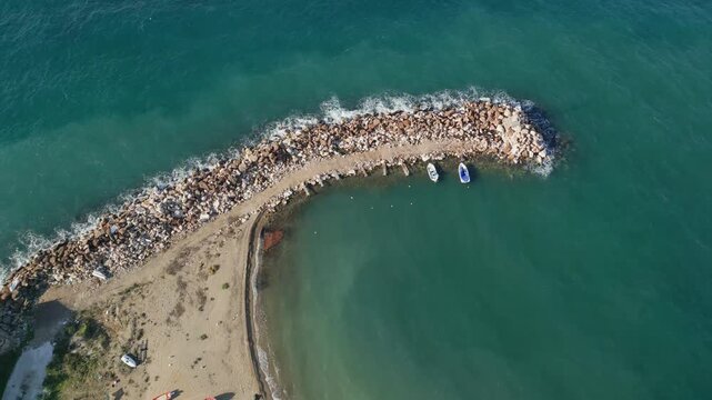 Aerial drone view of a curved rocky breakwater surrounded by clear turquoise sea water. Small boats anchored near the coast with gentle waves creating natural patterns