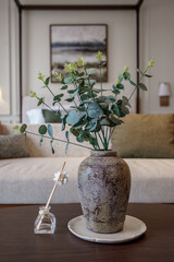 Artificial Eucalyptus plant in rustic vase and reed diffuser on table in cozy living room.