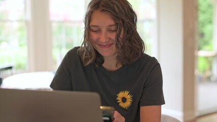 white woman smiling at phone and laptop, receiving positive message while wearing sunflower tshirt, joyful reaction and laughter, bright daylight, social connection and celebration mood